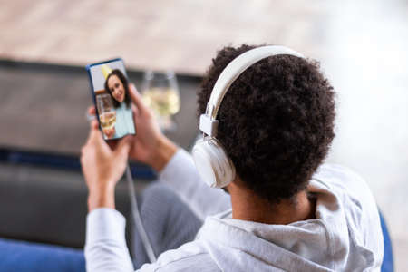 African american man is holding smartphone, chatting online with friend, sitting at home. Black guy...