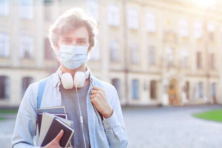 Young man in medical mask standing near closed for quarantine university campus.の写真素材