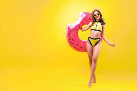 Happy young sexy woman with slim body wear bright yellow bikini swimsuit, hat and glasses is holding inflatable swimming donut ring isolated on orange background. Summer hotel pool sea rest concept.の写真素材