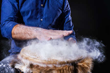 Man is playing on djembe drum, covered with talcum powder. Flour splashes on dark background. Summer festival concert performance. Ethnic rhythm. Percussion musical instruments and culture concept.の写真素材