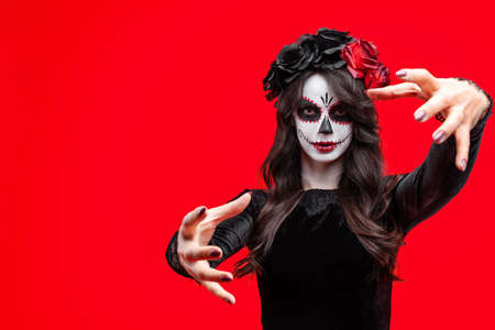 Girl with creative sugar skull makeup with a wreath of flowers on head, wide isolated on red...