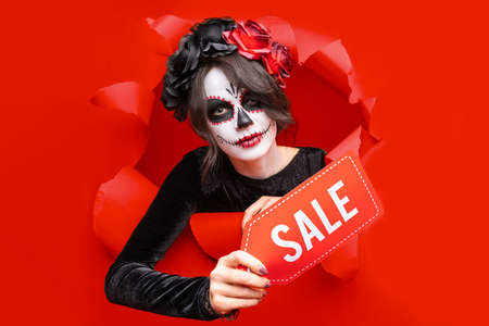 Girl with creative sugar skull makeup with wreath of flowers on head,  isolated red paper hole background. Concept Dia De Los Muertos poster for Halloween party or La Calavera Catrina. Holiday sale.の写真素材