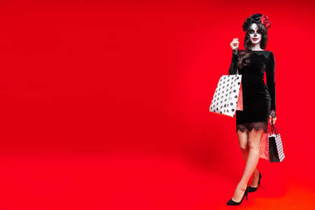 Girl with creative sugar skull makeup in wreath of flowers, isolated on red background hold shopping bags. Concept Dia De Los Muertos poster for Halloween party or La Calavera Catrina. Holiday sale.の写真素材