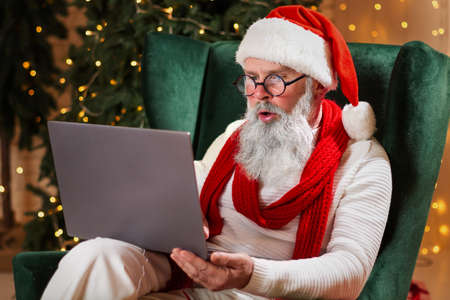 Serious Santa Claus sitting at his home and reading children's email letters , wish list on laptop near the fireplace and Christmas tree with gifts. New year and Merry Christmas happy holidays conceptの写真素材