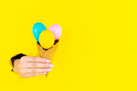 Abstract yellow background with female hand is holding three colorful eggs in ice cream cone. Minimal art design. Minimalist meal, fashion style. Festive food court. Happy Easter festival concept.の写真素材