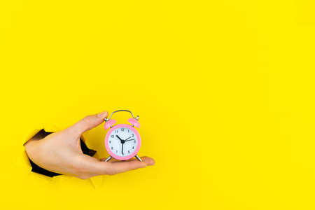 Ringing twin bell vintage classic pink alarm clock in female hand isolated in torn hole of yellow background. Rest hours time of life good morning night wake up awake concept. Mockup, copy space.の写真素材