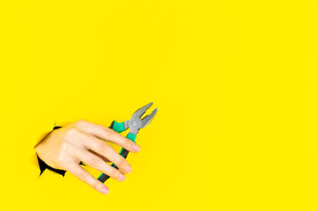 Female hand is holding tool wire cutters in torn hole of yellow background. Needle nose or pointy pliers for universal repair and construction. Copy space for text inscription. Building concept.の写真素材