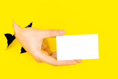 Female hand is holding, giving blank empty white paper card in torn yellow paper hole background. Breaking on, breakthrough. Ticket, flyer, invitation, greeting, coupon, template concept. Copyspace.の写真素材