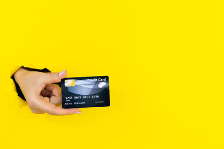 Closeup female hand is holding credit bank card in torn hole of yellow background. Template mockup with space for text. Online shopping, purchasing products concept. Pay by payment terminal.の写真素材