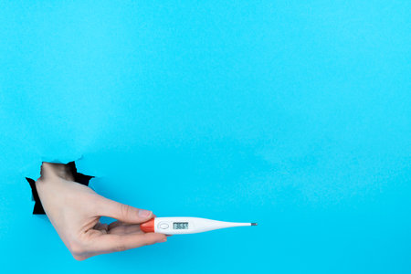 Female hand is holding electronic thermometer in torn hole of blue background. Medicine, health care...