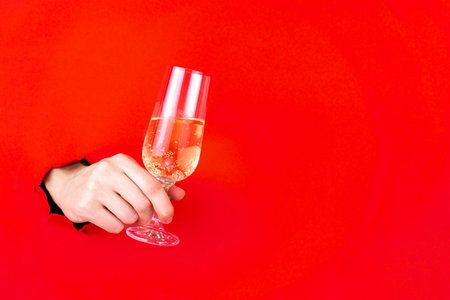 Female hand is holding glass of champagne in torn hole of red background. Christmas and New Year concept. Greeting festive invitation winter holiday card. Birthday or wedding. Copy space for text.の写真素材