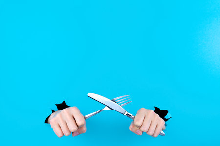 Female hands are holding cutlery metal knife and fork in torn hole of blue background. Food and restaurant menu concept. Serving dishes, lunch, waiting for order. Copy space for text inscription.の写真素材