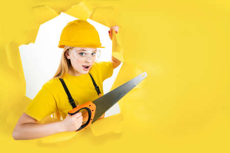 Energy fun handyman blond woman in protective helmet is holding manual saw in torn hole of yellow background. Female in male hard work. Renovation, repair, building concept. Emotional portrait.の写真素材