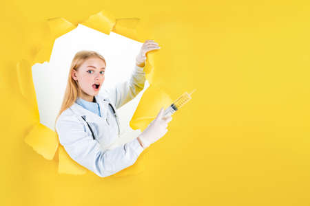 Young blond woman in white uniform in torn hole of yellow background. Female doctor is ready to patient visit. Professional therapist at work. Medicine, health care concept. Insurance card template.の写真素材