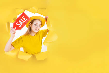Banner of blond trendy shopaholic woman excited about new purchases holding red paper sale label in torn hole of yellow background. Happy customer is ready for online shopping. Black friday concept.の写真素材