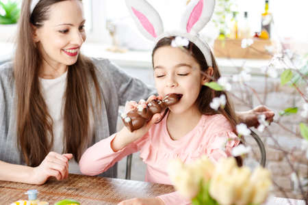 Close up happy mother daughter wearing bunny ears in kitchen during breakfast, eating delicious chocolate rabbit and colorful Easter eggs. Happy Easter atmosphere. Celebrating spring holiday concept.の写真素材