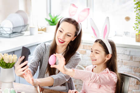 Mother and daughter doing selfie with Easter eggs, wearing bunny ears, have beauty and fun day...