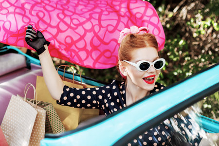 Beautiful stylish woman in vintage bright dotted clothes and glasses is driving mint old car after shopping. Fashion pin-up girl is waving pink scarf. Retro style concept. Black friday sale.の写真素材