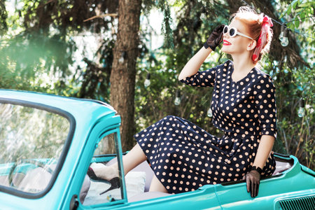 Beautiful stylish woman wearing vintage bright dotted clothes, sitting on mint old car hood. Fashion pin-up girl with makeup and hairstyle is walking outdoor. Retro style concept. Costume parties.の写真素材