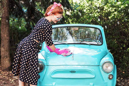 Beautiful stylish woman in vintage bright dotted clothes and glasses is washing mint old car....