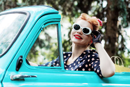 Beautiful stylish woman in vintage bright dotted clothes and glasses is driving mint old car after...