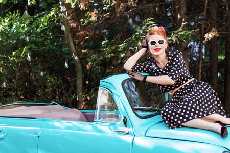 Beautiful stylish woman wearing vintage bright dotted clothes, sitting on mint old car hood. Fashion...