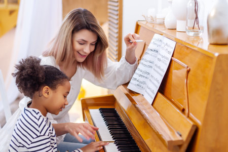 Woman and girl playing a piano. Beautiful mom teaching her daughter playing a piano. The piano teacher experienced pianist gives classes to a young little student. Hobby and activity for the children.の写真素材