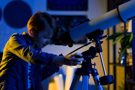 Boy carefully looks through a telescope. Smart inquisitive child explores sky through on a summer day with space maps on background. Cosmos and universe, Milky way stars. My astronomy science workの写真素材