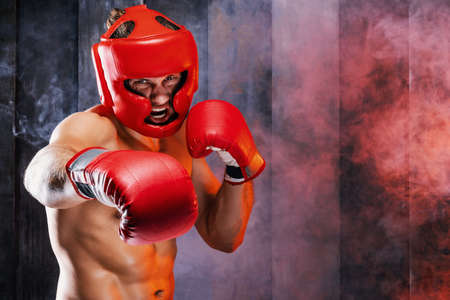 Dynamic motion portrait of sportive muscular athletic man in boxer gloves, helmet and shorts practicing attack. Fit strength male fighter kickboxing engaged in gym with screaming face in movement.の写真素材