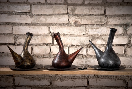 three old metallic jugs in front of brick wall