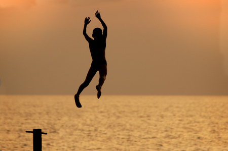 silouette of a boy jumping from pierce in front of brightly lighted seaの写真素材