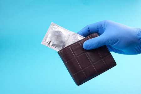 persons hand with medical gloves holds brown wallet. condoms at wallet over blue background