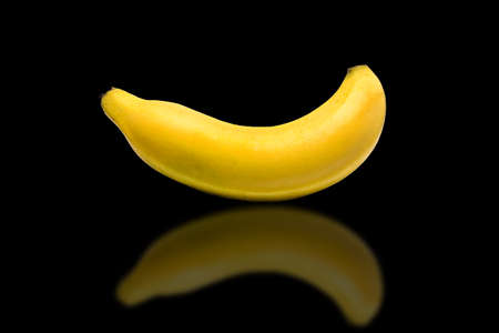 A single yellow banana laying on a black background with reflectionの写真素材