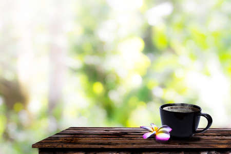 Black coffee cups and plumeria flowers on a wooden floor outside in the morning sun in a garden with beautiful bokeh.の写真素材