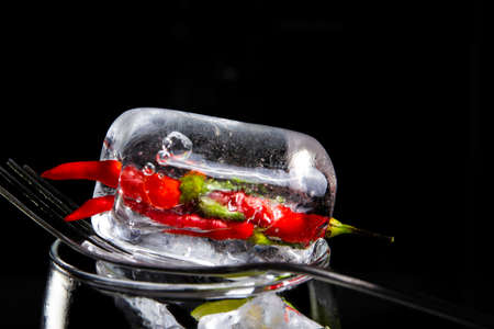 Ice cubes are placed on a spoon that rests on the mouth of a clear glass. Inside is wrapped in red and green peppers on a black background.の写真素材