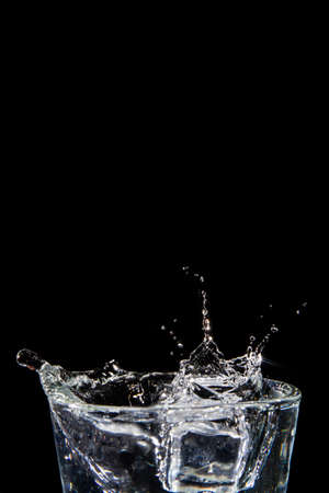 Water splashes caused by objects falling onto it. Water splashes on glass on isolated black background with close-up details. Side view of clear glass with liquid. Hand over the glass.の写真素材