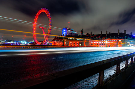 London Eye Ferris wheel in the City of London, illuminated at night with a long exposure, with lights on the line of cars and buses passing by at the time.のeditorial素材