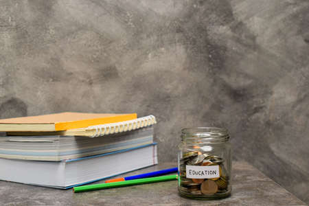 Coins in glass jar, book and pencil on marble table. Concept mone saving for education.