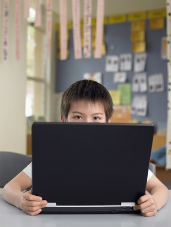 Elementary schoolboy sitting behind opened laptop in classroomの写真素材
