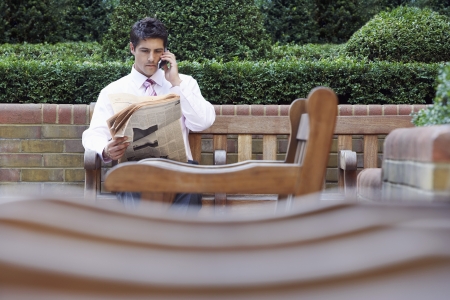 Businessman on park bench reading newspaper and talking on cell phoneの写真素材
