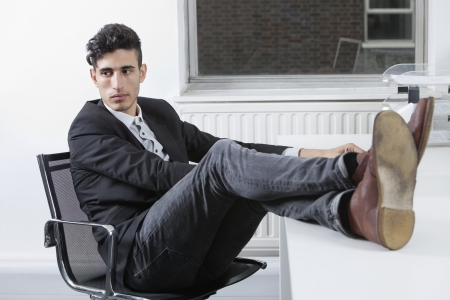 Thoughtful young businessman sitting with legs raised at desk in officeの写真素材