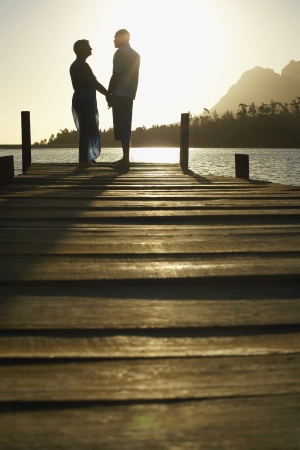 Couple standing on dock by lake holding hands side view.の写真素材