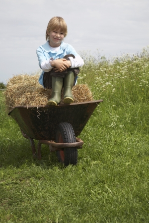 Boy (5-6) sitting on hay in wheelbarrow in field portraitの写真素材