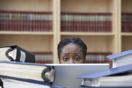Office worker using laptop sitting behind stacks of documents in office high sectionの写真素材