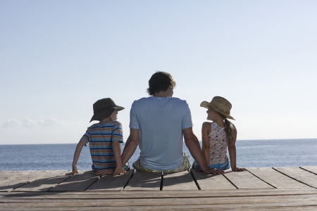 Father and two children (7-9) sitting on jetty back viewの写真素材