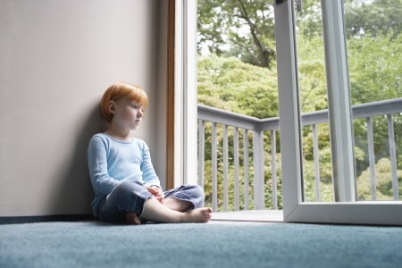 Young girl (5-6) sitting cross-legged on carpet looking through balcony doorの写真素材