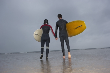 Couple of surfers carrying surfboards walking on beach back viewの写真素材