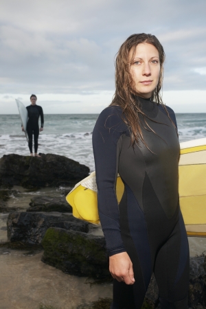 Female surfer carrying surfboard standing on beach another surfer standing in background portraitの写真素材