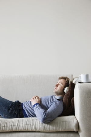 Man wearing headphones lying on sofa in living room side viewの写真素材