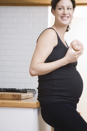 Pregnant woman eating doughnut in kitchenの写真素材